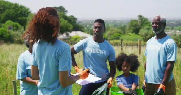 一个年轻的非洲裔美国妇女穿着蓝色T恤和橙色手套站在田里 拿着剪贴板 在收集垃圾和回收 慢动作之前指导各种各样的非洲裔美国志愿者的后视镜 — 图库视频影像