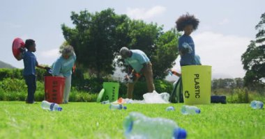 Farklı Afrika kökenli, yetişkin ve çocuklardan oluşan, üzerinde gönüllü yazılı eldiven ve tişört giyen, çöp toplayan ve geri dönüşüm yapan, yavaş çekim yapan bir grubun düşük açılı ön görüntüsü.