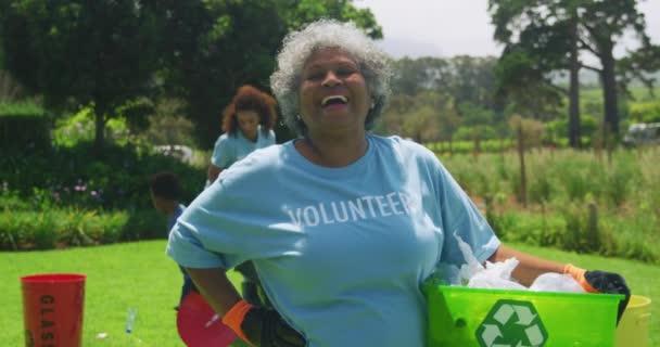 一位非洲裔美国老年妇女站在田里 戴着手套 穿着一件写有 志愿者 字样的T恤 手持绿色塑料回收箱 对着相机微笑 一群人在收集垃圾 这幅画像描绘了一位非洲裔美国老年妇女的形象 — 图库视频影像