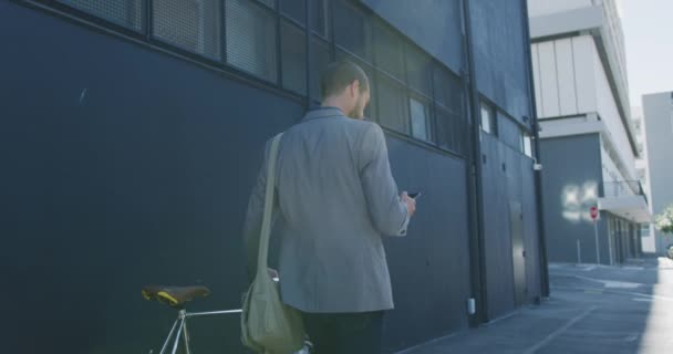 街中を自転車で歩き ビルが立ち並ぶ通りでスマートフォンを使用している若い白人実業家の背面図 スローモーション — ストック動画
