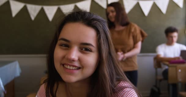 教室の机の上に座ってカメラに微笑む10代の白人女子高生の肖像画 背景で働く同級生 スローモーション — ストック動画