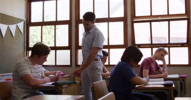 多民族の10代の男の子と女の子のクラスで机の間を歩く白人男性教師の正面図 彼らの仕事を見て スローモーション — ストック動画