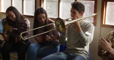 Sınıfta oturup müzik aletlerini çalan çok ırklı genç erkek ve kadın müzisyen grubunun ön görüntüsü, yavaş çekim.