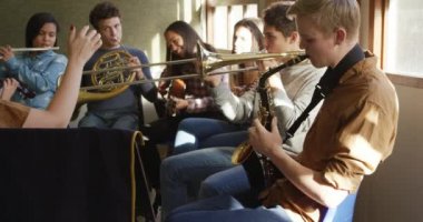 Bir okul bandosunda, ön planda beyaz bir kadın orkestra şefinin koluyla oynayan çok ırklı erkek ve bayan müzisyen grubunun yan görüntüsü, yavaş çekim.