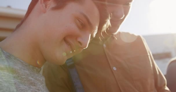 Vue de côté gros plan de deux adolescents garçons caucasiens utilisant une tablette et parlant dans leur terrain d'école, avec un groupe d'amis d'école parlant et utilisant des smartphones en arrière-plan, rétroéclairé par la lumière du soleil, ralenti 