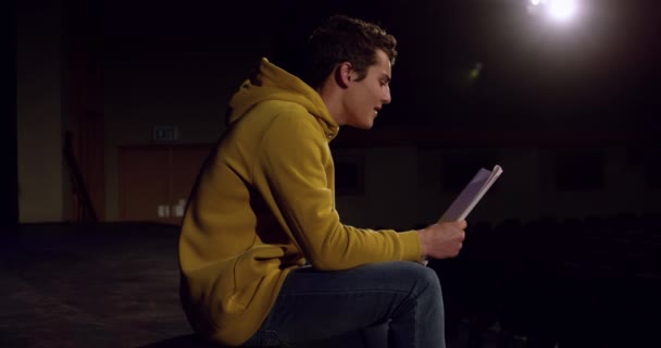 一个年轻的白人男孩坐在一个空荡荡的校园剧场的舞台上 正在为一场表演排练时从剧本中朗读 动作缓慢 — 图库视频影像
