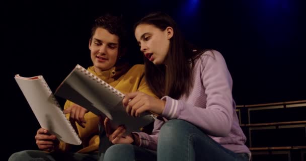 Seitenansicht eines kaukasischen Mädchens und Jungen im Teenageralter, die am Bühnenrand in einem leeren Schultheater sitzen, Drehbücher in der Hand halten und sich während der Proben für eine Aufführung anlächeln, Zeitlupe