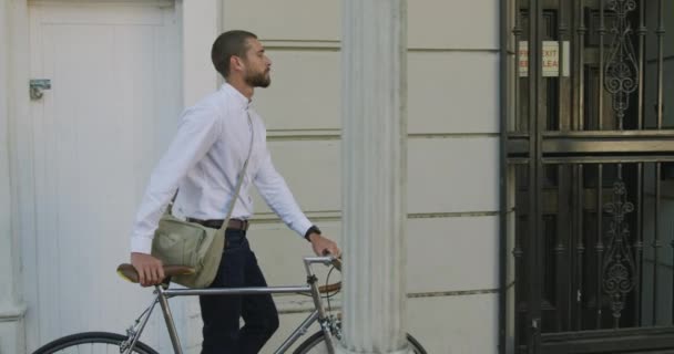 街を自転車で通りを歩いている若い白人実業家の側面図背景に建物がありスローモーション — ストック動画