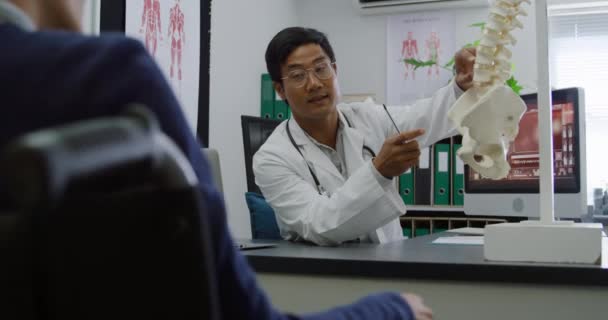 アジアの男性医師の話を聞き 彼の机の上の骨格のセクションを指して 病院のオフィスでの相談中に スローモーション4K — ストック動画