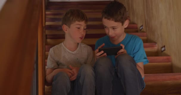 自宅の2人の若い白人の兄弟のフロントビューを閉じる 一緒にスマートフォンを使用して横に階段側に座って 話して スローモーション — ストック動画
