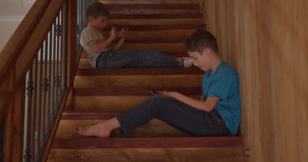 两个白人兄弟坐在家里的侧视图 他们坐在楼梯上 用智能手机 动作缓慢 — 图库视频影像