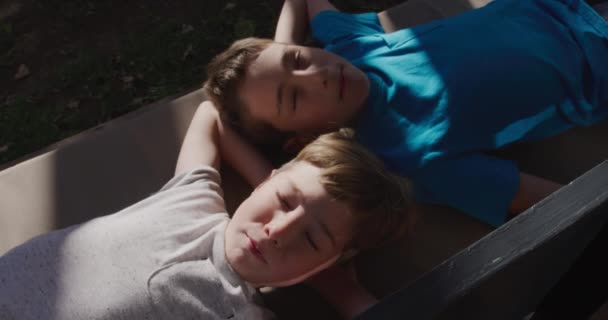 庭の自宅で2人の若い白人の兄弟の高い角度ビュー 彼らの頭と目の後ろに手で太陽の下でリラックスベンチシートの上に横たわっ閉じて スローモーション — ストック動画