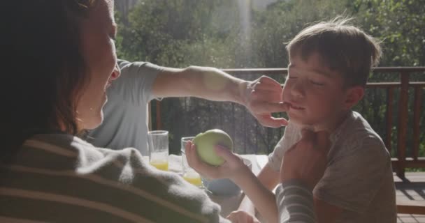 太陽のテーブルでテラスの外に座って 彼がリンゴを持っている間 両親は彼らの息子の顔をきれいに — ストック動画