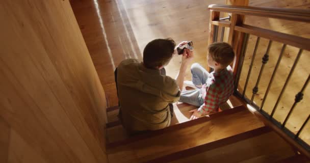 父が持っているスマートフォンを見て階段に座っている白人男性と自宅で彼の若い息子の高い角度のリアビュー スローモーション — ストック動画