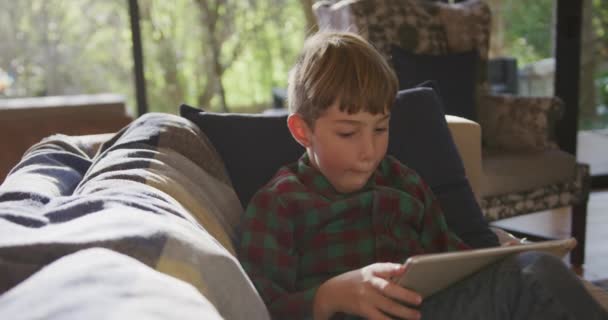 リビングの自宅で白人の少年のフロントビューを閉じると 太陽の下でソファの上にタブレットコンピュータを使用してリクライニング スローモーション — ストック動画
