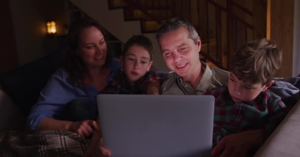 夜のリビングルームで二人の若い息子と一緒にソファに座ってノートパソコンを見て笑顔でゆっくりとした動き — ストック動画