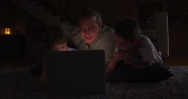 傍晚时分 一个白人男子和他的两个儿子躺在客厅里 躺在地板上 一起聊天 一起使用笔记本电脑 两个儿子躺在父亲的两边 动作缓慢 — 图库视频影像