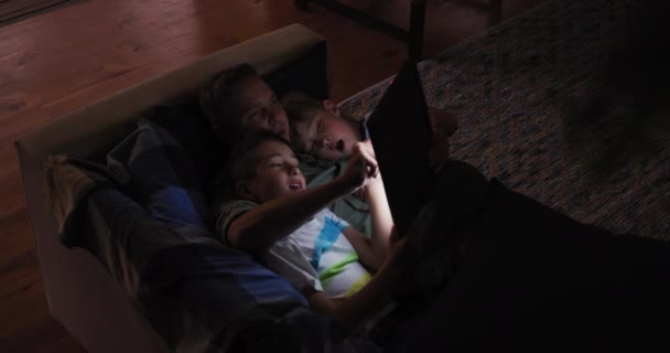 傍晚时分 一个白人男子和他的两个儿子坐在客厅里 躺在沙发上 一边聊天 一边看平板电脑 两个儿子把头靠在父亲胸前 动作缓慢 — 图库视频影像