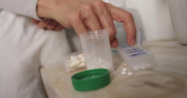 Evdeki yatağında yatan yaşlı beyaz bir kadının elini kapat, komodinine uzan ve diğer haplar ve komodinin üzerinde bir bardak suyla birlikte haftalık ilaç kutusundan bir tablet al, yavaş çekimde.