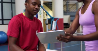 Spor salonundaki muayene masasında oturan iki protez bacaklı Afrikalı Amerikalı bir adamın yan görüntüsü. Spor için bacağını kontrol eden bir tablet bilgisayar kullanan Afrikalı Amerikalı bir kadın fizyoterapistle konuşuyor.