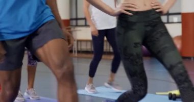 Beyazlı bir kadının spor merkezindeki fitness kursunda çeşitli yetişkinlerle egzersiz yaparken, ellerinin kalçasında ileri doğru hareketler yaparken yan görüntüsü.