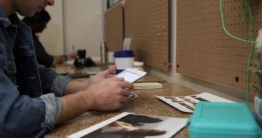 Yan bakışta, sıradan bir ofiste masada oturmuş tablet bilgisayar kullanan ve arka planda çalışan iki kadın meslektaşı olan genç bir erkek yaratıcı görülüyor.