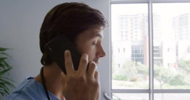 Kafkasyalı bir erkek sağlık görevlisi hastanede, merdivenlerde önlük giymiş, akıllı bir telefonla konuşuyor.