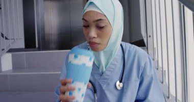 Önde, önlük, steteskop ve tesettür giyen, hastanede merdivenlerde oturan kahve içip pencereden dışarı bakan melez bir kadın var.