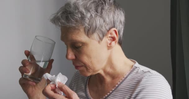 一位白发苍苍的老年妇女坐在旁边 手里拿着一杯水 在卧室里用纸巾擦拭她的鼻子 动作缓慢 — 图库视频影像