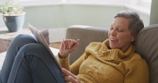 ビデオ通話 手と笑顔 スローモーションのためのタブレットコンピュータを使用して彼女のリビングルームのソファに座って 自宅で上級白人女性のフロントビュー — ストック動画