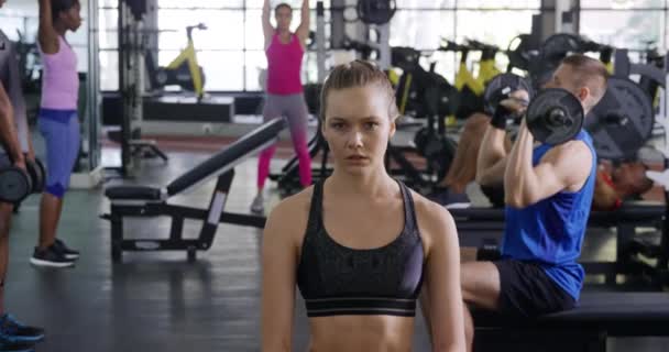 一名身穿运动服的年轻白人女运动员的画像 她在健身房训练后的休息中期待着拍照 背景上有一群不同的成年人在使用体育用品 动作缓慢 — 图库视频影像
