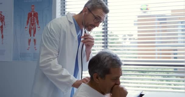 一名白种人男医生戴着眼镜 身穿实验室外套 在医院里与一位年龄较大的混血男性患者交谈 背靠听诊器听他呼吸的侧视图 — 图库视频影像