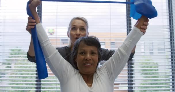 白人女性理学療法士のフロントビューは 混合人種の女性患者の後ろに立って 彼女は病院での理学療法セッション中に抵抗バンドで彼女の腕を行使するのに役立ちます — ストック動画