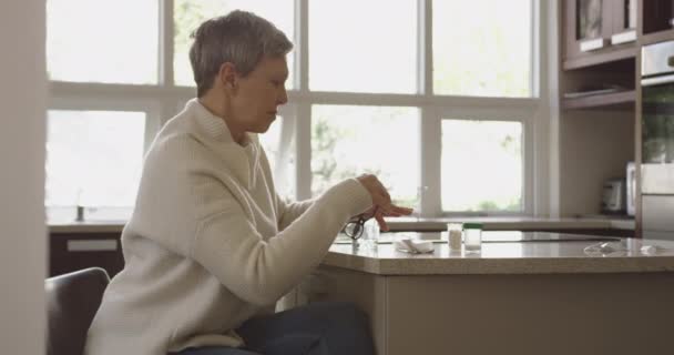 一位白种人老妇人的侧影 她一头灰白的短发 坐在厨房的工作台上 戴上眼镜 阅读药物 面前放着药瓶和一杯水 动作缓慢 — 图库视频影像