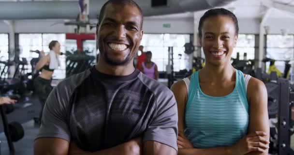 アフリカ系アメリカ人の男性と 体育館でのトレーニングの合間にカメラを見ながら笑顔でスポーツ服を着た混合人種の女性の肖像 背景にあるジム機器を使用した多様な大人のグループ スローモーション — ストック動画
