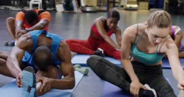 在体育中心的健身课上 一群身穿运动服的成年人坐在练习垫上 前倾着抓住一条伸出的腿的脚踝 站在那里观看 — 图库视频影像