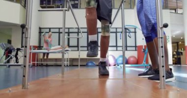Spor salonunda spor kıyafetleri giymiş, önünde protez bacakları olan Afrikalı Amerikalı bir adamın önünde diz çöküp paralel barlarda yürümesine yardım eden Afrikalı Amerikalı bir kadın fizyoterapist.