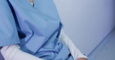 Bir hastane ameliyathanesinde önlük giyen, duvara yaslanmış, ağır çekimde yüzünü çeviren melez bir kadın sağlık uzmanının yan görüntüsü.