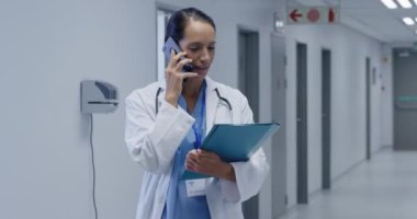 Kafkasyalı, laboratuvar önlüğü ve steteskop giymiş mutlu bir kadın doktorun hastane koridorunda dikilip akıllı bir telefonla konuştuğunu ve elinde bir pano tuttuğunu görmek.