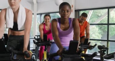 Spor salonunda spor kıyafetleri giyen ve spor bisikletleri üzerinde bisiklet süren bir grup yetişkinin ön görüntüsü.