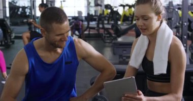 Spor salonundaki eğitimden sonra spor kıyafetleri giyen beyaz bir erkek ve kadının ön görüntüsünde boynunda bir havlu var ve adama ağır çekimde bir tablet bilgisayar gösteriyor.