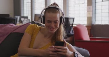 Modern bir ofiste, koltukta oturmuş kulaklıkla müzik dinleyen, akıllı telefonuyla mesaj atan, ağır çekimde gülümseyen, yaratıcı, profesyonel bir kadının ön görüntüsü.