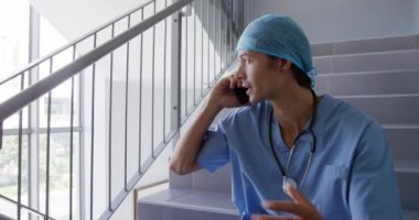 Kafkasyalı bir erkek sağlık görevlisinin hastanede, ameliyat önlüğü ve kepiyle merdivenlerde otururken, akıllı bir telefonla konuşurken, kalkıp merdivenlerden inerken,