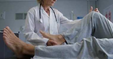 Laboratuvar önlüğü giymiş beyaz bir kadın doktor bacağını ağır çekimde hareket ettirirken, son sınıf melez bir erkek hastanın kanepeye uzanması.