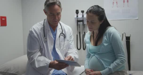 研究室のコートと聴診器を身につけ妊婦の白人女性患者と話をし診察中にタブレットコンピュータを見せました — ストック動画