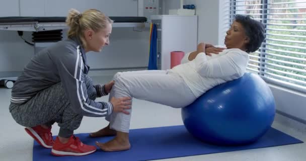 一位白种人女性理疗师帮助一位混血女性病人进行运动的侧视图 她在医院的理疗期间靠在一个运动球上站稳了脚 — 图库视频影像
