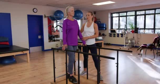 一名白人老年妇女在体育中心体育馆与一名白人女性理疗师一起锻炼的侧视图 引导她通过一个锻炼项目 走在平行的酒吧之间 — 图库视频影像