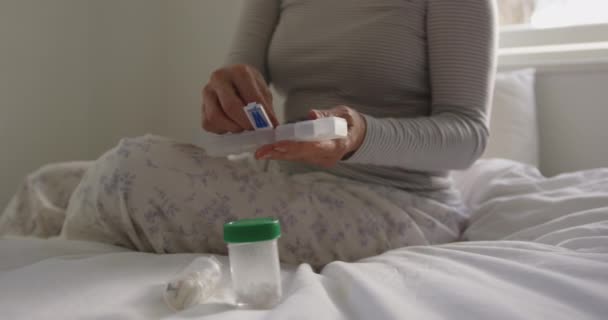 一位年长的白人妇女坐在自己家里的床上 从每周的药箱里拿出药丸 拿在手里 身边还拿着其他药箱 慢吞吞地躺在床上 — 图库视频影像