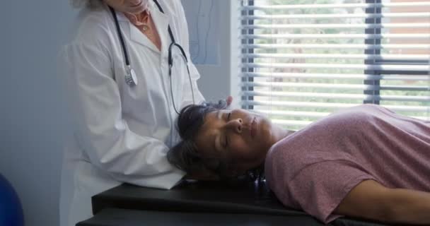一名身穿实验室外套的白人女医生在一家医院的理疗过程中把头抬起来 在沙发上躺着一位混血女病人的侧影 — 图库视频影像