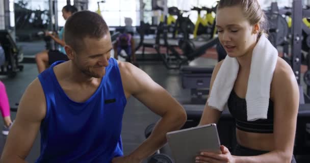 体育館でトレーニング中の休憩中にスポーツ服を着た白人男性と女性の正面図は 彼女の首の周りにタオルを持っており 男にタブレットコンピュータ スローモーションを示しています — ストック動画